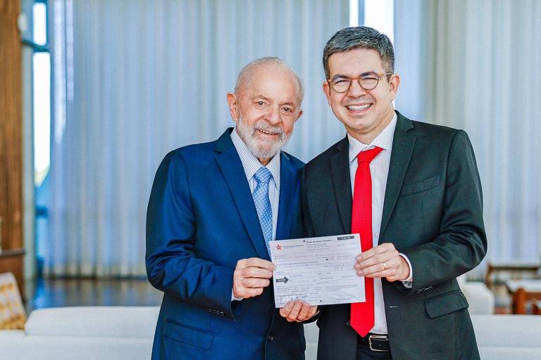 Na foto é possível ver dois homens de pé segurando juntos um papel; Lula está à esquerda da imagem, de azul, e Randolfe à direita, de vermelho