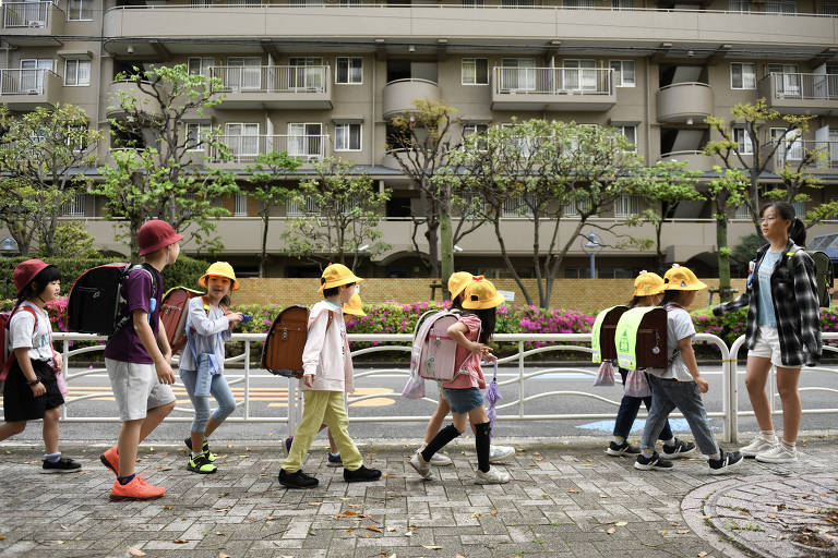 A imagem mostra um grupo de crianças caminhando em uma calçada, acompanhadas por uma mulher adulta. As crianças estão usando mochilas grandes e chapéus amarelos, exceto duas que usam chapéus vermelhos. Elas parecem estar indo para a escola. Ao fundo, há um prédio residencial com várias varandas e árvores podadas ao longo da calçada.
