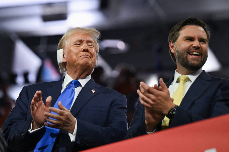 A imagem mostra dois homens de terno aplaudindo em um evento. O homem à esquerda é Donald Trump, que está usando um terno azul escuro com uma gravata azul e o homem à direita é J.D. Vance, que está usando um terno escuro com uma gravata amarela. Ambos parecem estar em um ambiente interno com iluminação artificial.
