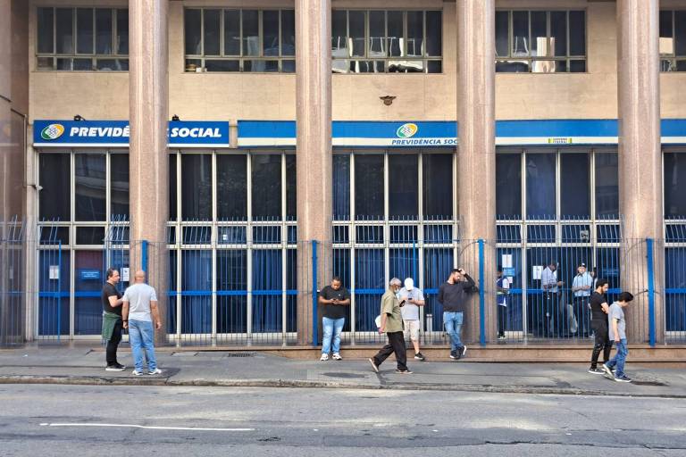 Imagem de uma fachada de agência da Previdência Social com pessoas caminhando na calçada em frente. A agência possui grandes janelas de vidro e colunas verticais