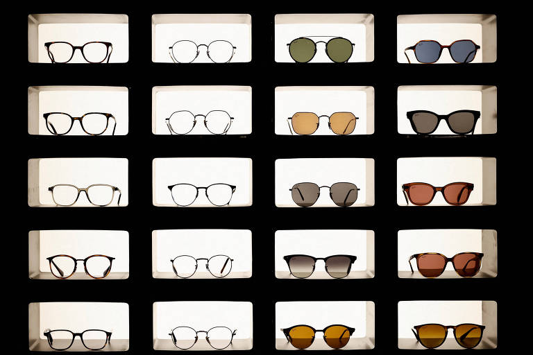 A imagem mostra uma exposição de óculos em uma vitrine iluminada. Há cinco fileiras e quatro colunas de compartimentos, cada um contendo um par de óculos. Os óculos variam em estilo, cor e formato, incluindo armações redondas, quadradas e aviador, com lentes de diferentes tonalidades.