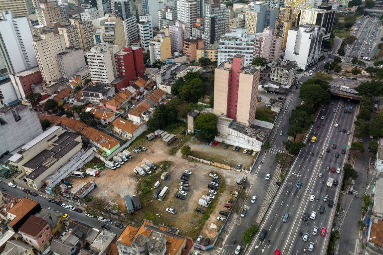 Imagem aérea mostra terreno ao centro com carros estacionado com mato alto em meio a muitos prédios e uma grande avenida à direita