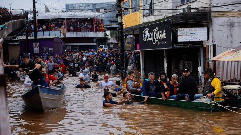 A imagem mostra uma rua inundada com várias pessoas sendo resgatadas em barcos. A água marrom cobre a rua, e há uma grande multidão de pessoas ao fundo, algumas em áreas mais elevadas. Vários indivíduos estão dentro da água, ajudando no resgate