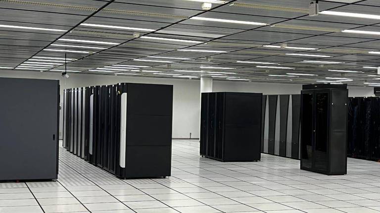A imagem mostra um centro de dados com várias fileiras de racks de servidores pretos organizados em um ambiente limpo e bem iluminado. O teto possui iluminação fluorescente e o piso é composto por placas brancas, provavelmente elevadas para facilitar a ventilação e o cabeamento