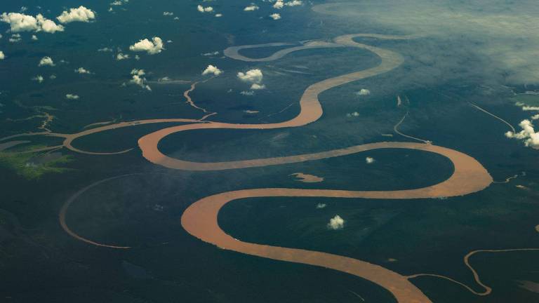 Imagem aérea de um rio sinuoso com águas marrons, serpenteando através de uma vasta área verde. Pequenas nuvens brancas estão espalhadas pelo céu acima do rio