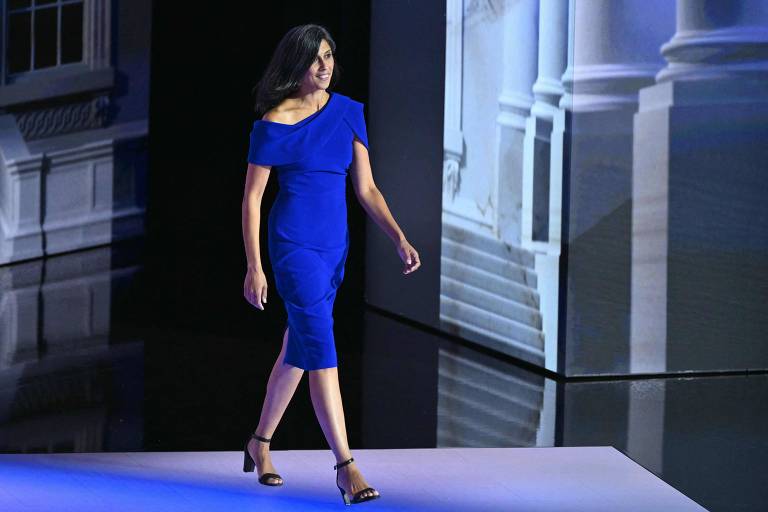 Uma mulher está desfilando em uma passarela. Ela veste um vestido azul de um ombro só e sandálias de salto alto pretas. O fundo da passarela tem uma imagem projetada de uma escadaria e colunas arquitetônicas.