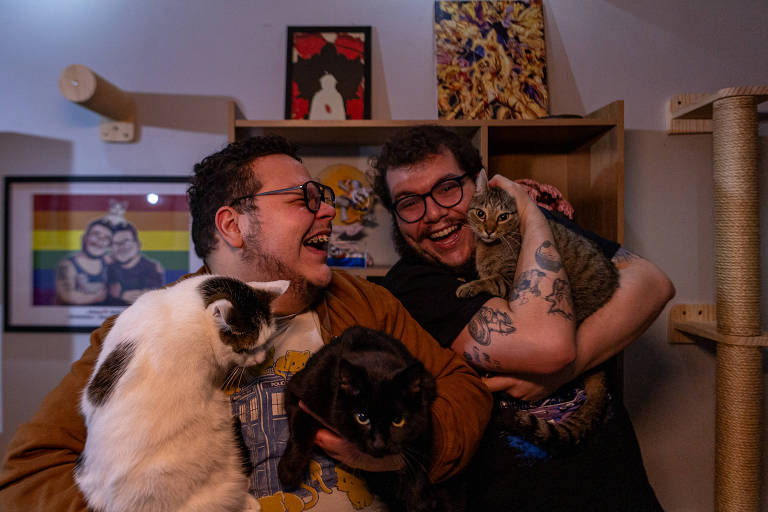 A imagem mostra duas pessoas sorrindo e segurando três gatos. Uma pessoa está à esquerda, segurando um gato branco e preto e um gato preto. A outra pessoa está à direita, segurando um gato cinza. Ao fundo, há uma parede com uma bandeira do arco-íris e várias obras de arte penduradas. Há também uma estrutura de madeira para gatos.
