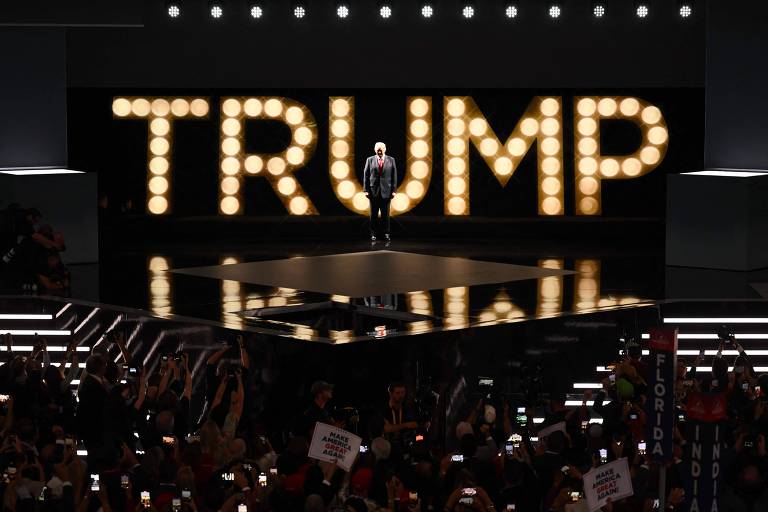 Homem de terno escuro e gravata vermelha à frente da palavra Trump, escrita com luzes