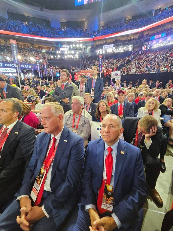A imagem mostra um grande grupo de pessoas sentadas em um evento formal, possivelmente uma conferência ou convenção. Muitas pessoas estão vestindo ternos e gravatas vermelhas. O ambiente é um auditório ou arena com iluminação brilhante e várias bandeiras ao fundo. Há uma atmosfera de formalidade e atenção.