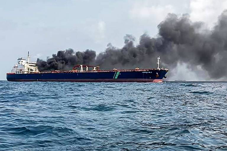 A imagem mostra um navio-tanque no mar com uma grande quantidade de fumaça preta saindo de sua parte traseira. O céu está claro e o mar está calmo. A fumaça indica que há um incêndio a bordo do navio.