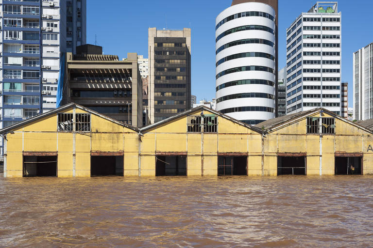 A imagem mostra um cenário de inundação em uma área urbana, com um edifício amarelo parcialmente submerso em água marrom. Ao fundo, há vários prédios altos, incluindo um com uma fachada listrada em branco e preto. O céu está claro e azul, contrastando com a situação de alagamento.