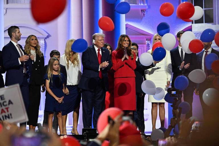 Um grupo de pessoas está em um palco decorado, cercado por balões vermelhos, brancos e azuis. No centro, um homem de terno escuro e gravata vermelha está ao lado de uma mulher vestida de vermelho. Outras pessoas ao redor estão vestidas em trajes formais, e há uma criança de vestido azul. No canto inferior esquerdo, há um cartaz com a inscrição 'MAKE AMERICA GREAT AGAIN'.