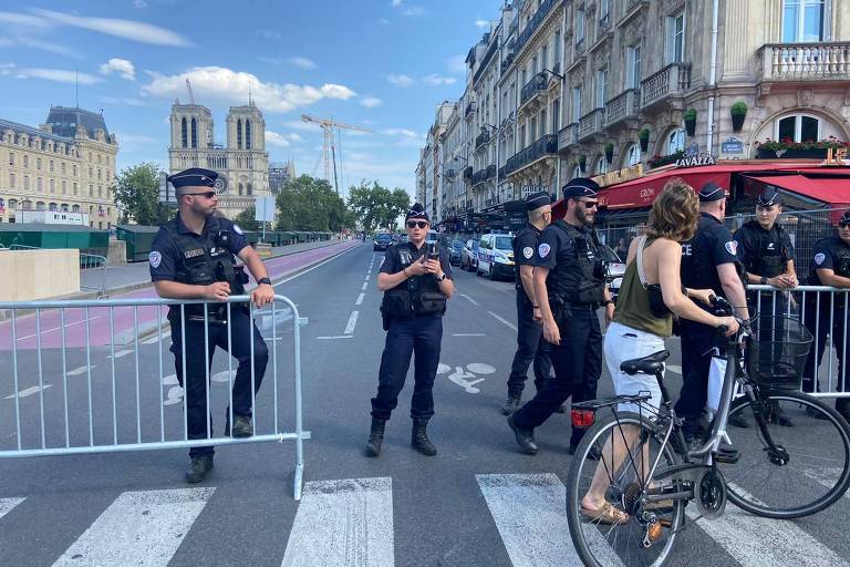 A imagem mostra vários policiais em uma rua bloqueada por barreiras de metal. Alguns policiais estão de pé, enquanto outros estão conversando. À direita, uma mulher está andando de bicicleta. Ao fundo, há edifícios históricos e um céu azul com algumas nuvens.