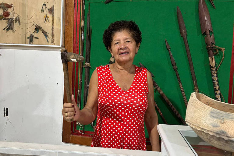 Uma mulher de vestido vermelho com bolinhas brancas está em um museu. Atrás dela, há uma parede verde com várias lanças e outros artefatos indígenas pendurados. À esquerda, há quadros com ilustrações de figuras humanas. Em primeiro plano, há vitrines brancas exibindo outros objetos.
