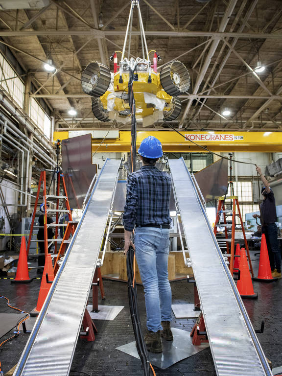 A imagem mostra um trabalhador de costas, usando um capacete azul e uma camisa xadrez, em uma fábrica. Ele está observando um equipamento de elevação que está suspenso no ar. O ambiente é industrial, com várias estruturas metálicas, cones de segurança laranja e uma grua amarela no fundo.
