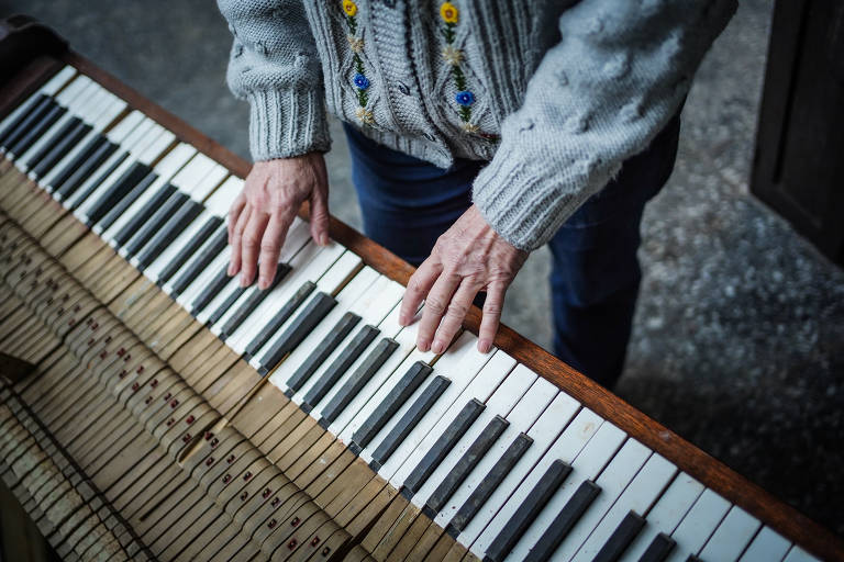 A imagem mostra uma pessoa usando um suéter cinza com detalhes coloridos, tocando um piano. O piano parece estar desgastado, com algumas teclas danificadas ou faltando. A pessoa está de pé, com as mãos sobre as teclas do piano.