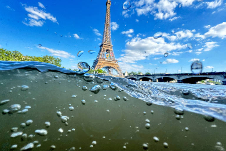 A imagem mostra uma vista única da Torre Eiffel em Paris, capturada parcialmente debaixo d'água. A parte inferior da imagem está submersa, com bolhas visíveis na água, enquanto a parte superior mostra a Torre Eiffel contra um céu azul com algumas nuvens brancas. Ao fundo, há uma ponte sobre o rio Sena.