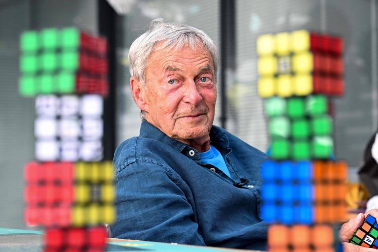 Um homem idoso está sentado, vestindo uma camisa azul, com uma expressão serena. Em primeiro plano, há várias torres de cubos mágicos (Rubik's Cubes) de diferentes cores, desfocados, que enquadram a imagem.