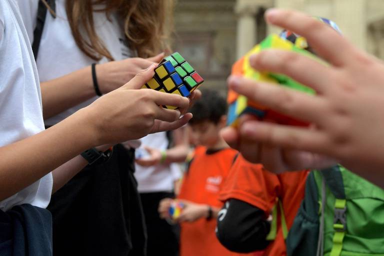 A imagem mostra várias pessoas segurando e resolvendo cubos mágicos (Rubik's Cubes). As mãos de várias pessoas são visíveis, cada uma manipulando um cubo colorido. Ao fundo, outras pessoas também estão envolvidas na atividade
