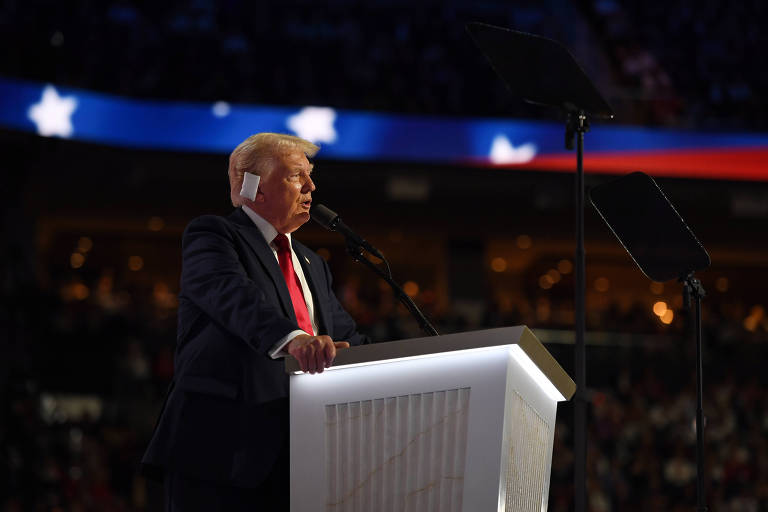 A imagem mostra Donald Trump, um homem de terno escuro e gravata vermelha discursando em um palco, com parte da orelha enfaixada. Ele está atrás de um púlpito branco com um microfone, e há um teleprompter à sua frente. Ao fundo, há uma faixa com estrelas brancas e cores azul e vermelha, sugerindo um evento patriótico ou político.
