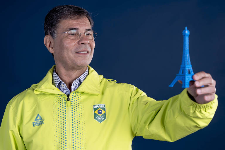 Um homem de óculos está segurando uma miniatura azul da Torre Eiffel. Ele veste uma jaqueta amarela com o logotipo da marca 'Peak' e o emblema do Comitê Olímpico Brasileiro, que inclui a palavra 'Brasil' e os anéis olímpicos. O fundo é azul escuro.
