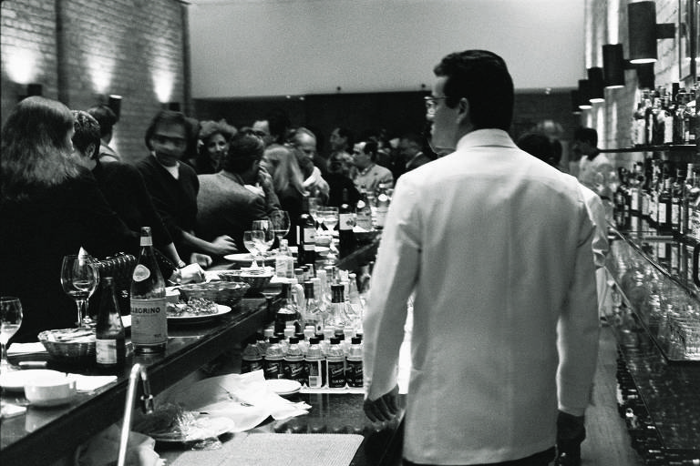 Foto em preto e branco; em primeiro plano, vemos um garçom vestido com um terno branco. À esquerda, há o balcão do restaurante, cheio de pratos e copos. Pessoas estão sentadas conversando