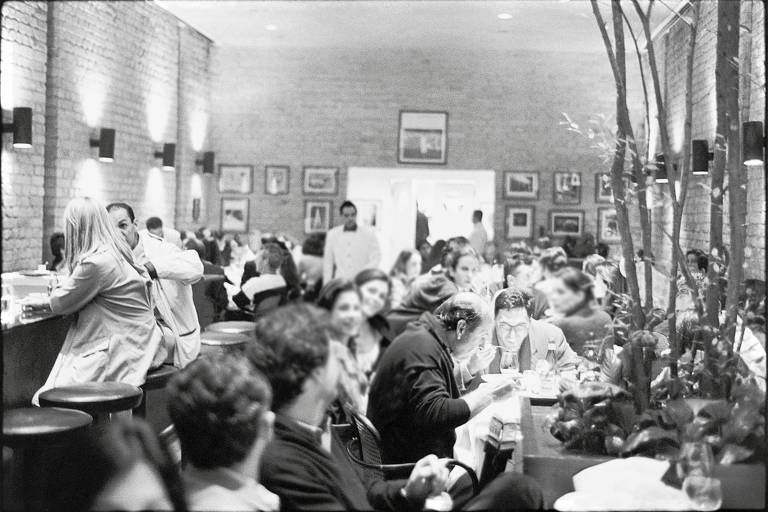 Ambiente interno do restaurante, com paredes de tijolões. Todas as mesas estão lotadas, com pessoas conversando