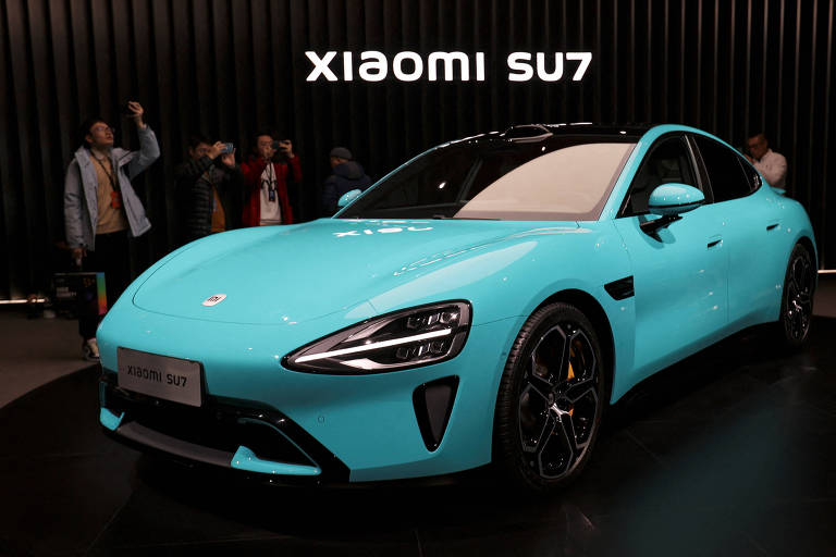 Imagem de um carro azul-claro, modelo Xiaomi SU7, em exposição. O carro possui um design moderno e aerodinâmico, com faróis elegantes e rodas esportivas. Ao fundo, há várias pessoas observando o veículo e uma parede preta com o texto 'XIAOMI SU7' em branco.