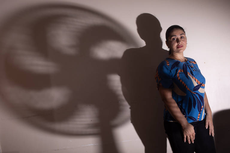 Imagem mostra uma mulher de pé ao lado de uma parede, com uma sombra projetada atrás dela. A sombra tem formato de um ventilador. A mulher está usando uma blusa azul com detalhes coloridos e calça preta