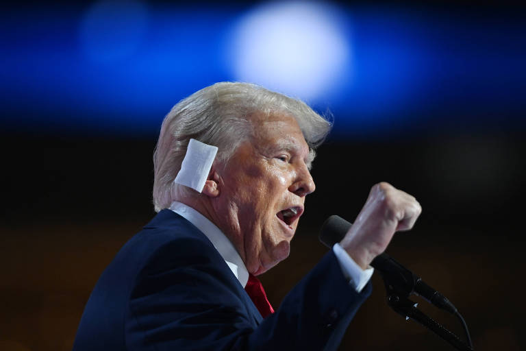 Donald Trump, homem branco, pele rosada, está em frente a um microfone, em uma tribuna, onde faz um discurso. Está de perfil para a câmera, com a mão direita à altura do rosto. Tem cabelos brancos e um curativo na orelha direita. Usa terno preto, camisa branca e gravata vermelha 