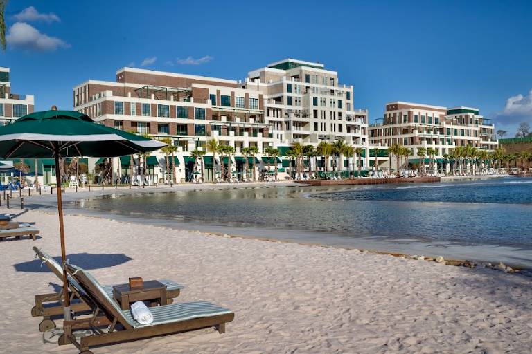 A imagem mostra uma praia com areia clara e cadeiras de praia sob guarda-sóis verdes. Ao fundo, há um grande resort com vários andares e varandas, situado à beira-mar. O céu está claro e azul, sugerindo um dia ensolarado.