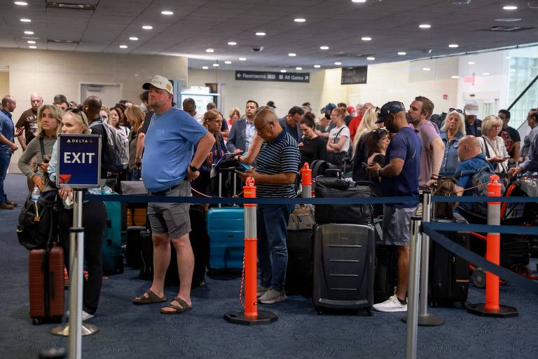 Imagem de uma fila de passageiros em um aeroporto. As pessoas estão em pé, aguardando em uma área delimitada por barreiras e cones laranja. Algumas pessoas estão segurando ou ao lado de suas malas. Há um sinal que indica 'EXIT' à esquerda. O ambiente é iluminado e parece movimentado, com várias pessoas ao fundo.