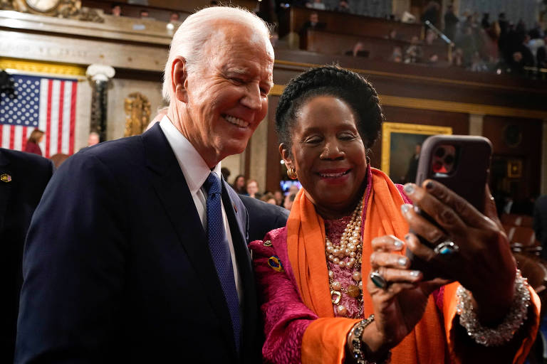 A imagem mostra duas pessoas sorrindo enquanto tiram uma selfie. A pessoa à esquerda é Joe Biden, um homem branco, com cabelos brancos, que está vestido com um terno escuro e gravata, enquanto a pessoa à direita é a deputada Sheila Jackson Lee, uma mulher negra, com cabelos pretos, que usa um traje colorido com um xale laranja. Ao fundo, há uma bandeira dos Estados Unidos.
