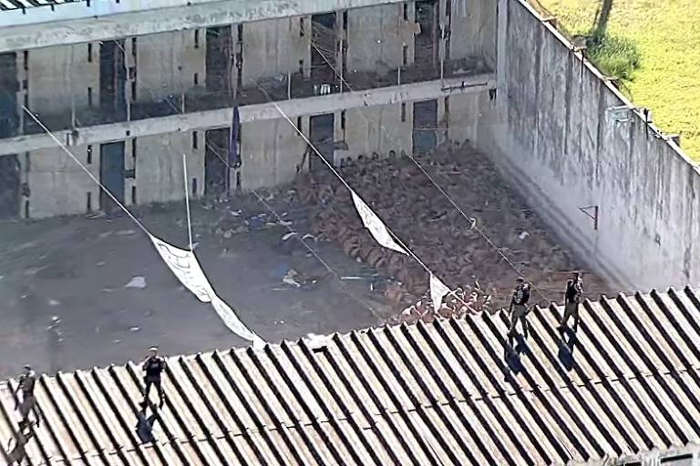 Foto aérea mostra um grupo de presos sentados no chão e com as cabeças abaixadas. eles estão agrupados em um canto de um pátio, perto do muro. no telhado, na parte inferior da imagem, policiais armados observam a cena. ao fundo estão portas de diversas celas em dois pavimentos