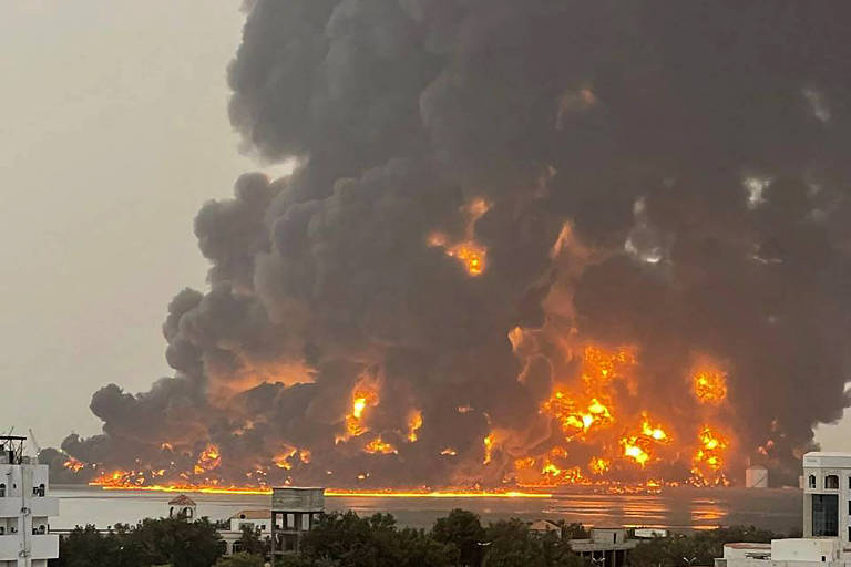 Grande incêndio, com fogo e colunas de fumaça, em uma região à margem do mar. Foto foi tirada de uma outra margem, onde há alguns edifícios