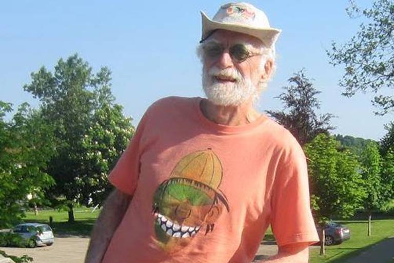 Um homem idoso com barba branca e óculos de sol está sorrindo ao ar livre. Ele usa um chapéu branco e uma camiseta laranja com uma estampa de um rosto sorridente. Ao fundo, há árvores verdes e um céu azul claro