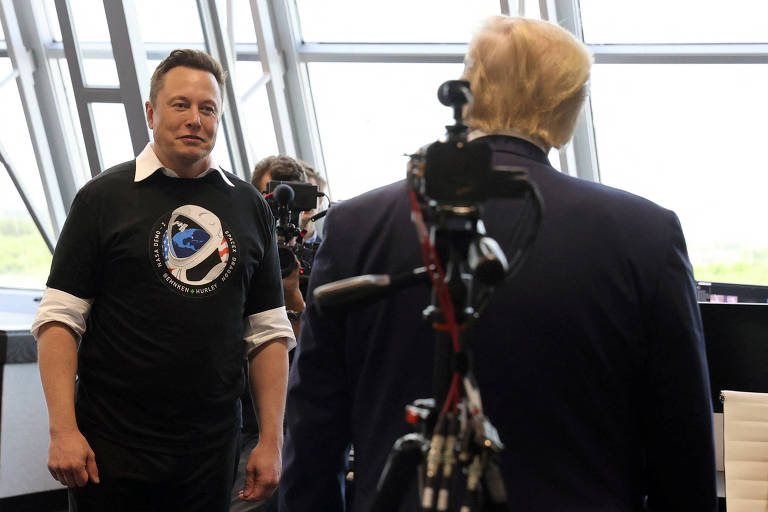 À esquerda, Elon Musk, de camisa branca sob moletom preto, caminha rumo a Donald Trump, à direita, que está de costa e usa paletó escuro.