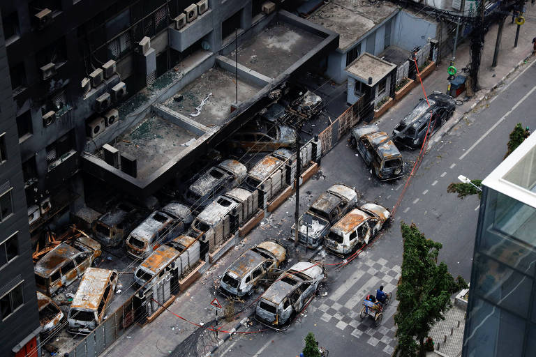 Imagem aérea de uma rua urbana com vários veículos queimados e danificados estacionados ao longo da calçada. Os edifícios ao redor também mostram sinais de danos, com fachadas escurecidas e janelas quebradas. A rua está parcialmente bloqueada e há poucos pedestres visíveis.