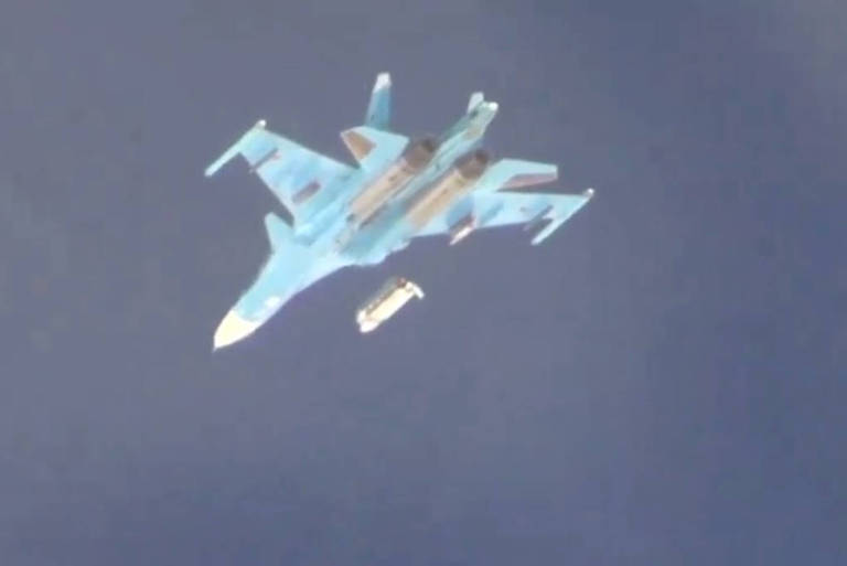 Avião militar azul lança uma bomba branca. O céu está azul