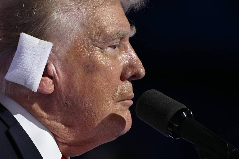 A imagem mostra Trump, um homem branco com cabelos brancos, de perfil, com um curativo branco na orelha. Ele está falando em um microfone preto. O fundo da imagem é escuro.
