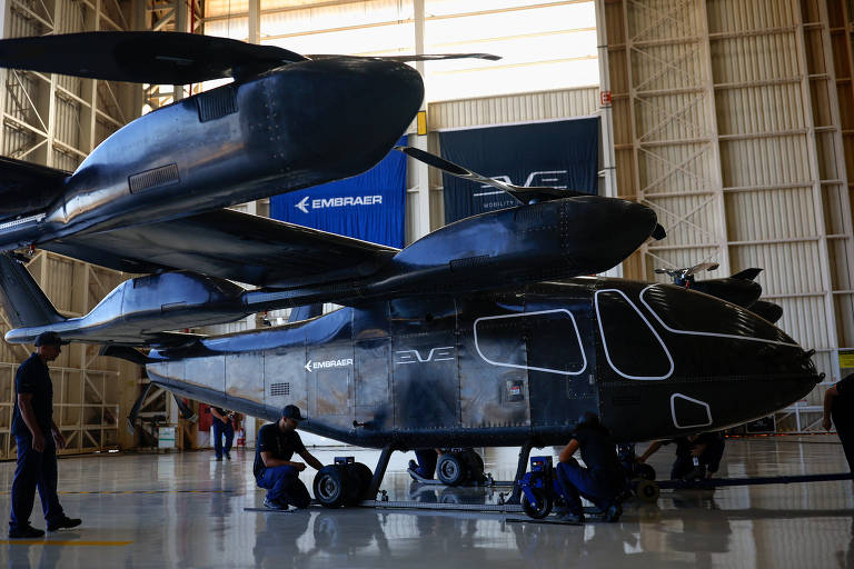 Imagem de um helicóptero preto estacionado dentro de um hangar. Há vários técnicos ao redor da aeronave, aparentemente realizando manutenção ou inspeção. O hangar é espaçoso e bem iluminado, com estruturas metálicas visíveis ao fundo.