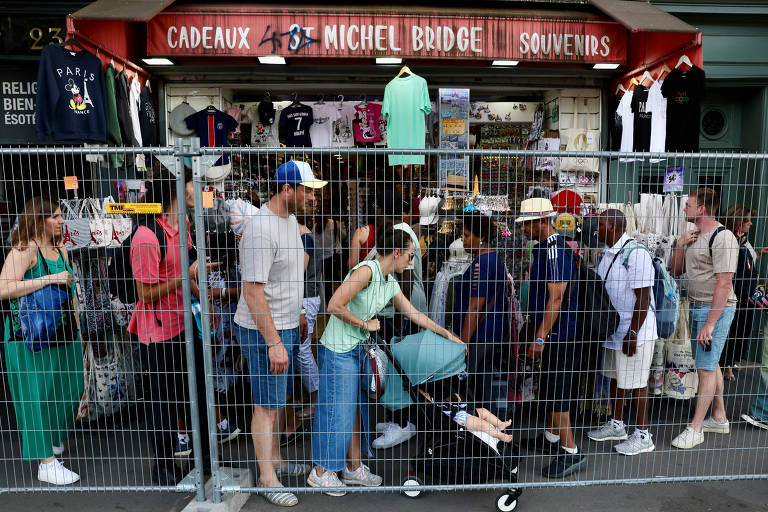 Imagem mostra uma fila de pessoas em frente a uma loja de souvenirs chamada 'Michel Bridge Souvenirs'. A loja vende diversos itens, incluindo camisetas, bolsas e outros produtos turísticos. As pessoas estão alinhadas atrás de uma cerca de metal. A fachada da loja é vermelha e há várias camisetas penduradas na entrada.