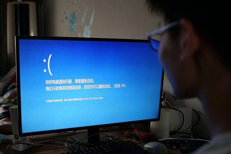 Um homem olha para a tela do computador, que exibe uma tela azul com um emoji de rosto triste. Trata-se de uma tela de erro indicando que o computador travou, com texto em chinês Imagens como esta foram significativamente menos frequentes na China do que no resto do mundo.