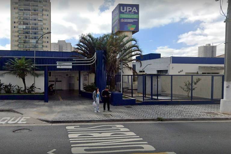 A imagem mostra a entrada de uma Unidade de Pronto Atendimento em São Bernardo do Campo. A fachada é azul e branca, com uma placa grande no topo que diz UPA Baeta Neves Unidade de Pronto Atendimento 24 horas'