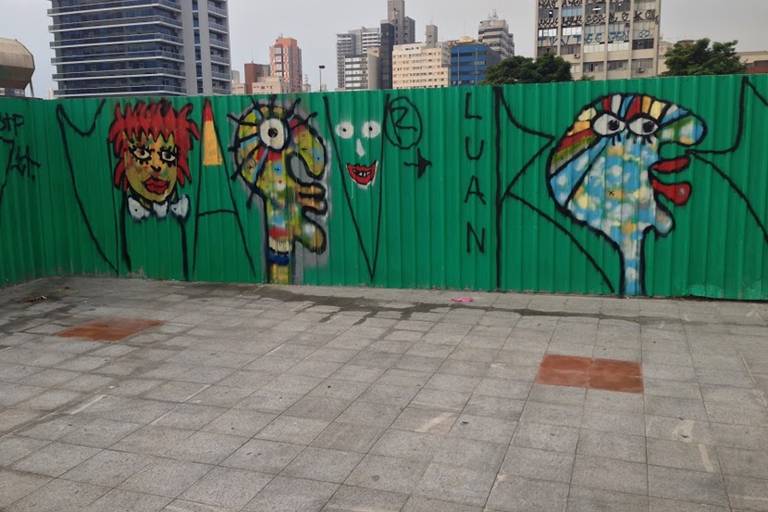 A imagem mostra uma cerca verde com murais de arte urbana pintados. Há três figuras principais: uma com cabelo vermelho e rosto amarelo, outra com um corpo verde e olhos grandes, e a terceira com um rosto azul e cabelo colorido. Ao fundo, é possível ver prédios altos e um céu nublado