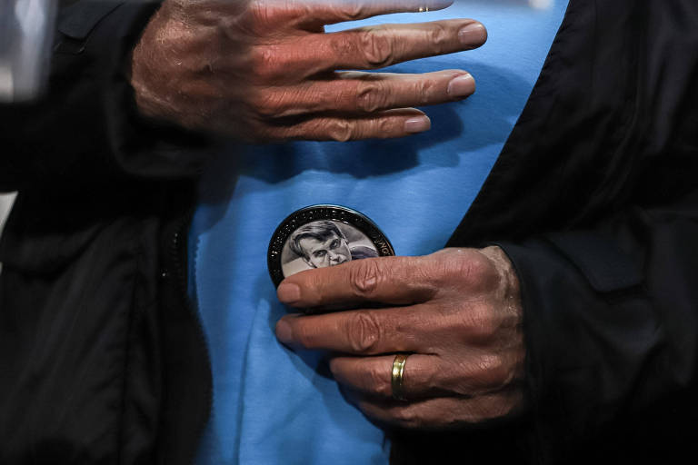 A imagem mostra as mãos de Jair Bolsonaro segurando uma medalha. A mão esquerda está posicionada acima da medalha, enquanto a mão direita a segura por baixo. Ele está vestindo uma roupa preta e usa uma aliança dourada no dedo anelar da mão direita