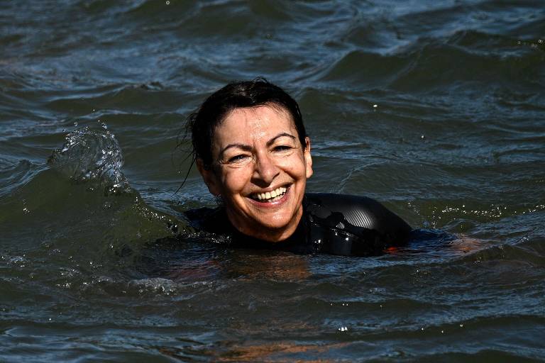 Uma mulher sorridente está nadando em águas do mar. Ela usa um traje de neoprene preto e parece estar se divertindo. O sol reflete na água.