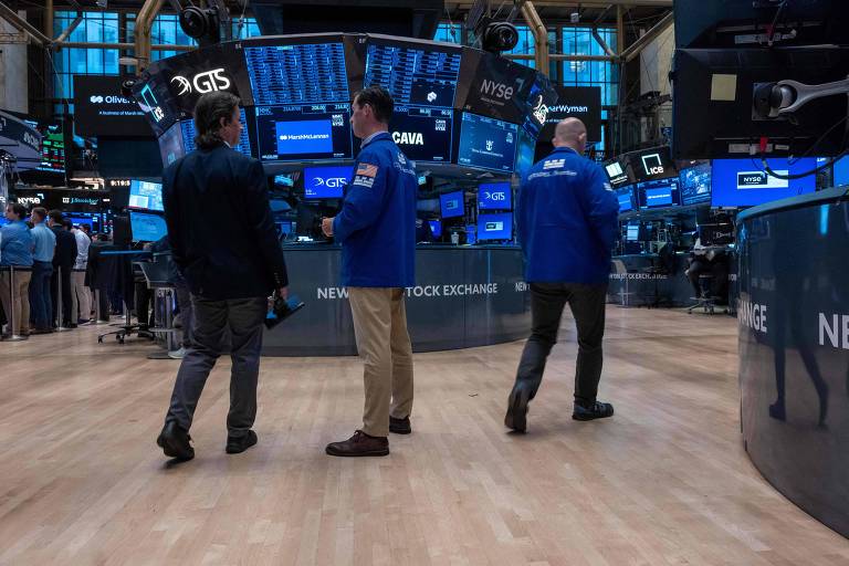 A imagem mostra três pessoas de costas, vestindo jaquetas azuis, no piso de negociação de uma bolsa de valores. Ao fundo, há várias telas exibindo gráficos e informações financeiras. O ambiente é iluminado e há outras pessoas ao redor, também envolvidas em atividades relacionadas ao mercado financeiro.
