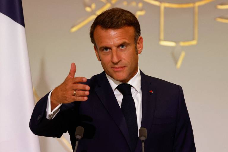 Imagem de um homem de terno escuro e gravata, fazendo um gesto com a mão direita durante um discurso. Ele está em frente a um microfone, com uma bandeira parcialmente visível à esquerda e um símbolo dourado no fundo