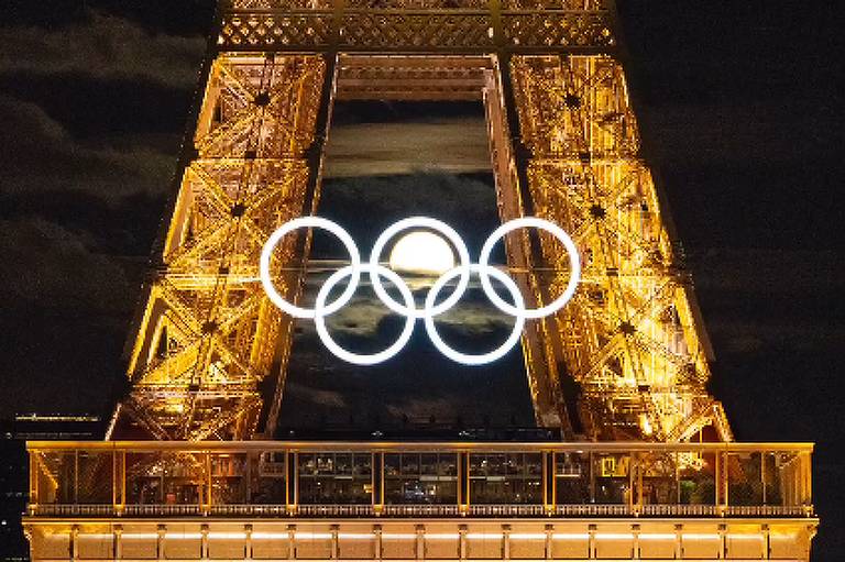 A imagem mostra a Torre Eiffel iluminada à noite, com os anéis olímpicos brilhando no centro da estrutura. No canto superior direito, há um pequeno logotipo dos anéis olímpicos. A iluminação dourada da torre contrasta com o céu noturno escuro.